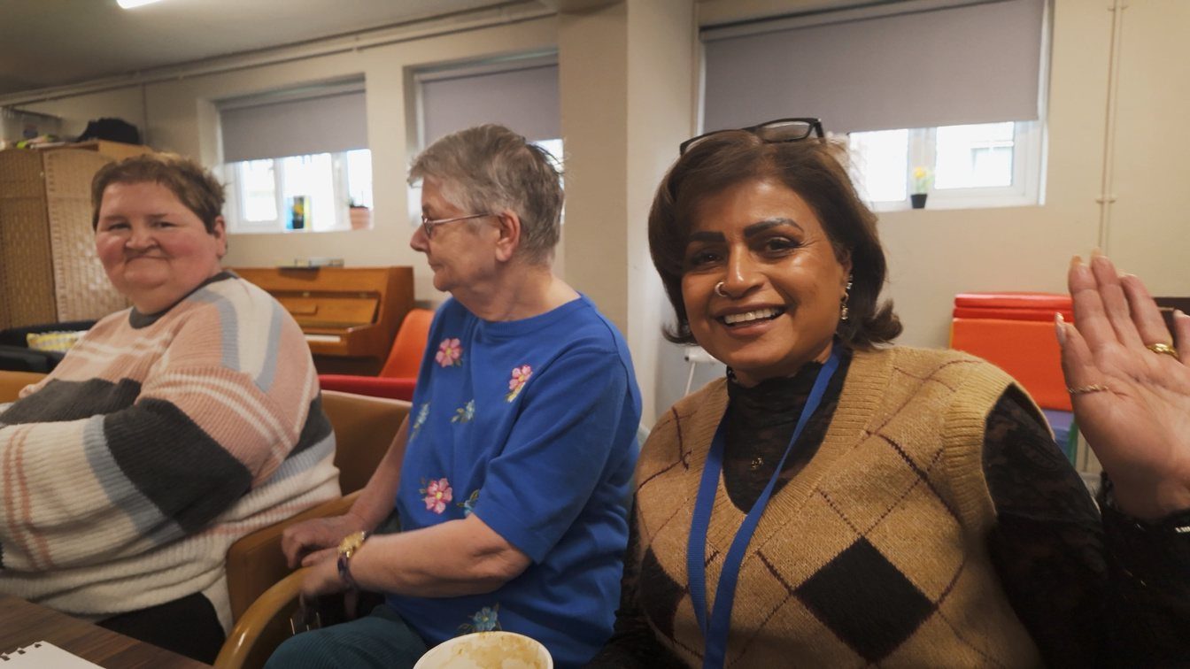 Three women laughing and waving at a table in a Barking and Dagenham community centre